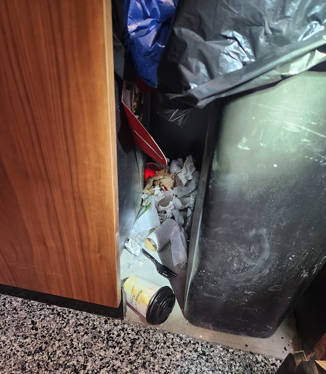 Trash and spilled food debris piled behind a garbage bin inside a New Jersey office building, creating a hidden sanitation issue that attracts house flies and other nuisance flies.